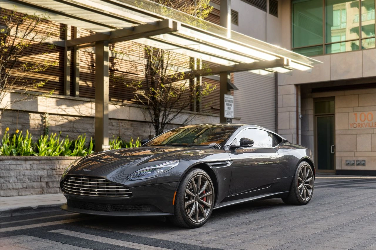 Black Aston Martin DB12 sports car parked under architectural pergola with modern building and landscaping
