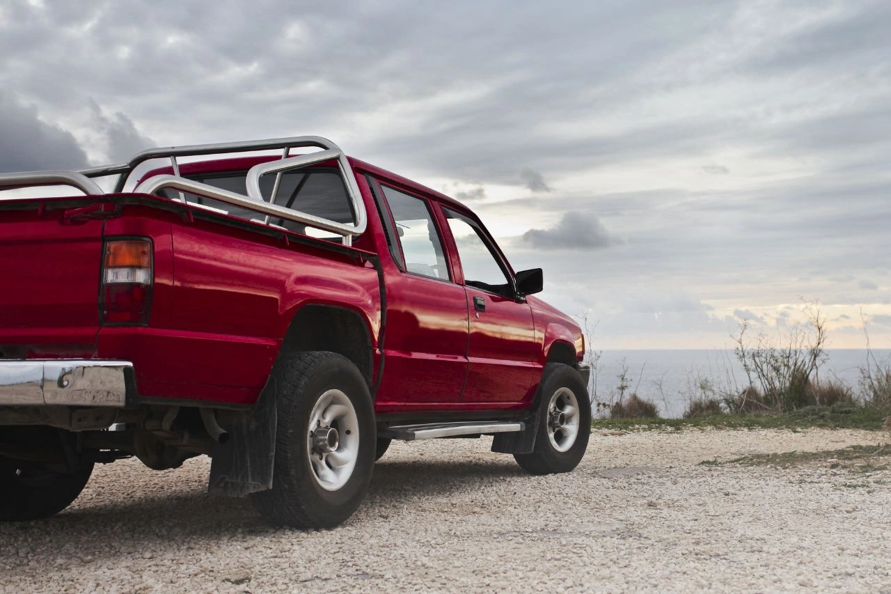 Red pickup truck with roll bar parked on sandy terrain with cloudy sky background